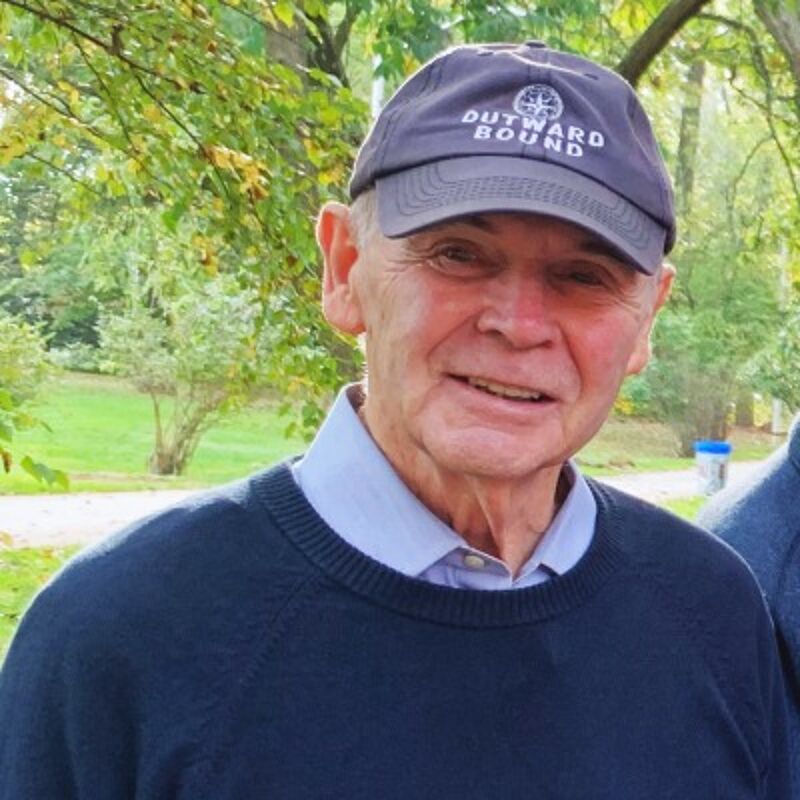 The image shows an older man wearing a navy blue cap with the words "OUTWARD BOUND" on it. He is also wearing a navy blue sweater over a light blue collared shirt. The man is smiling and appears to be outdoors, with trees and greenery in the background.